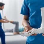 Podiatrist showing anatomical foot skeleton model during his assistant examination injured leg of patient in department of traumatology and orthopedics
